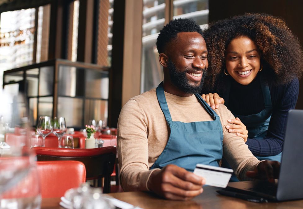 Two small business owners smiling while using a laptop and credit card in their restaurant, representing financial growth and community lending success.